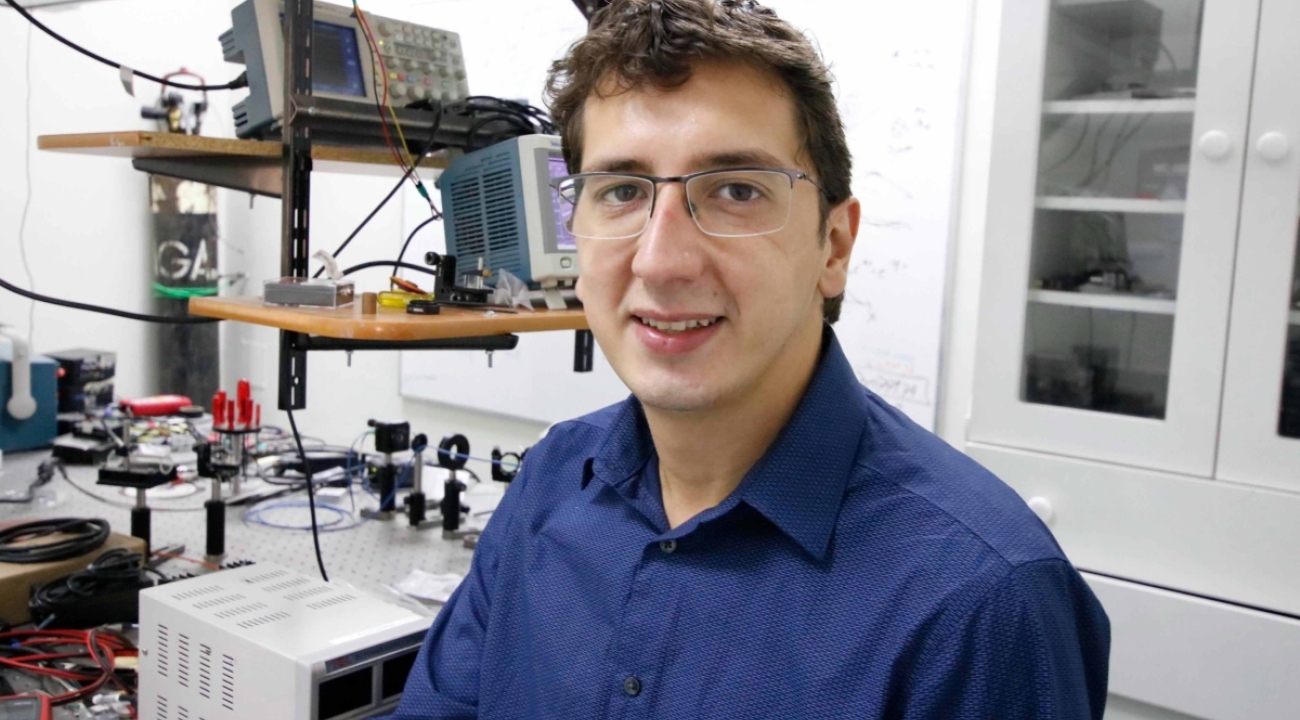 Man in a blue button up shirt in front of various electrical and optical pieces of lab equipment.