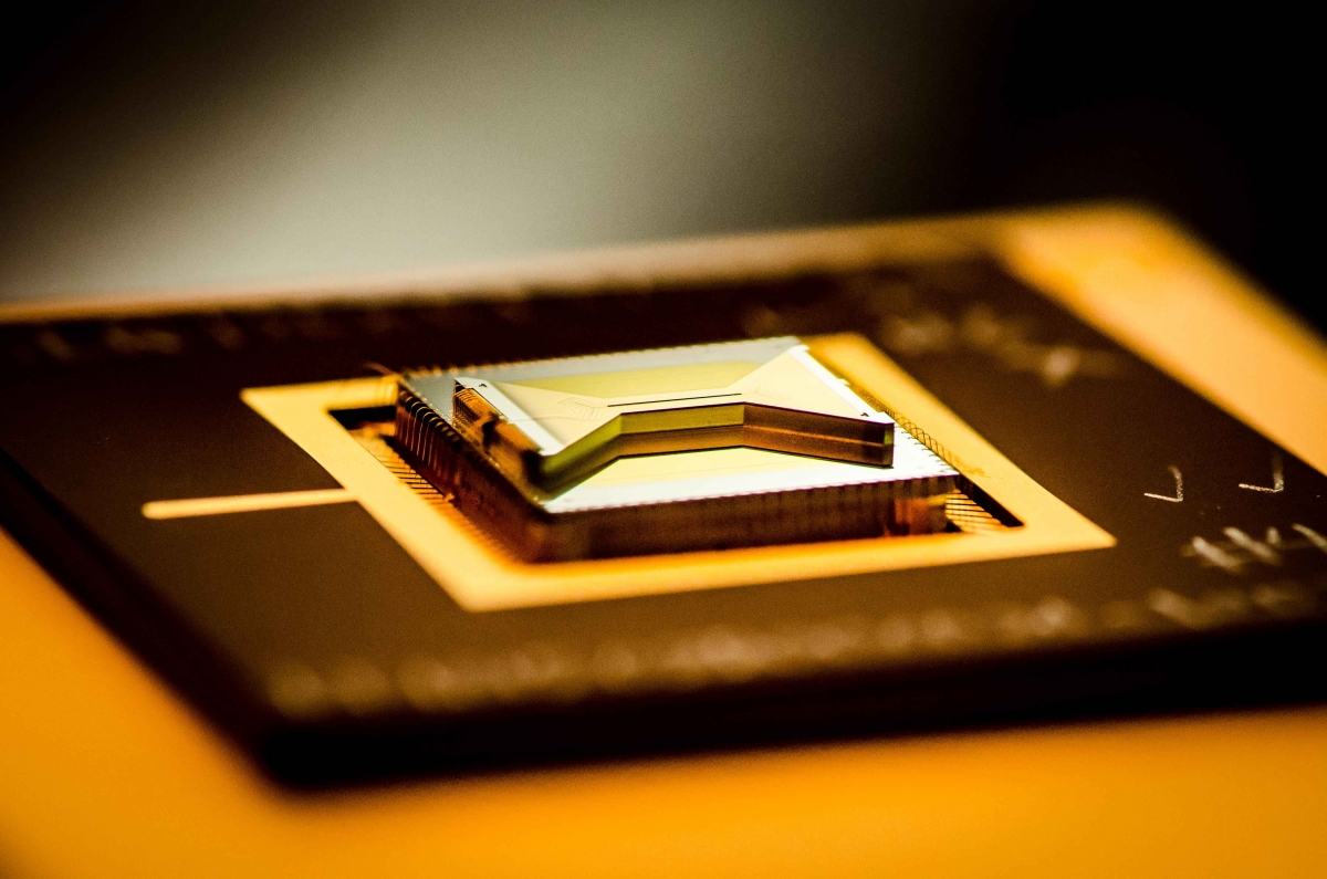 A chip made of golden bow-tie-shaped structure on top of a dark rectangular base that is used to contain ions for experiments and quantum computing tasks. The base of the chip has illegible markings on it.