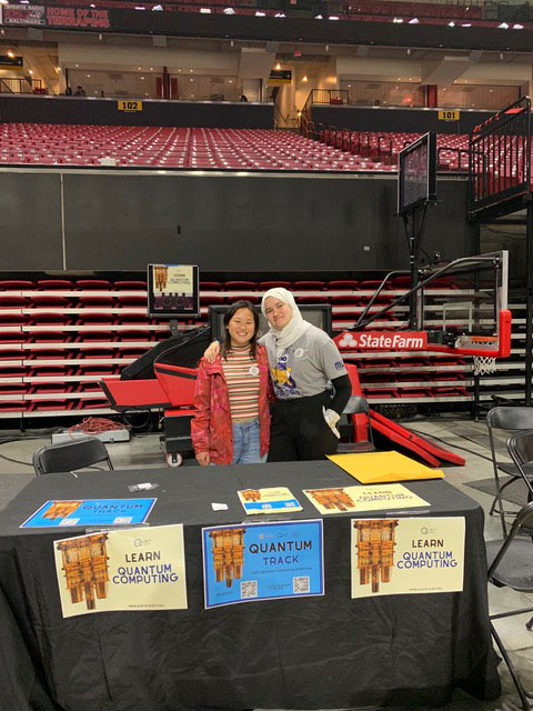 Two woman stand behind a table covered with a black table cloth and signs. They are in front of indoor basketball bleachers.