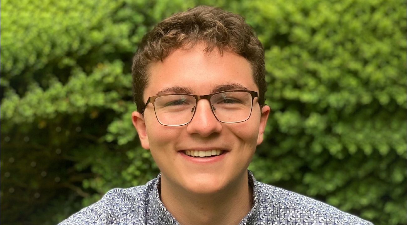 A man wearing glasses stands in front of green shrubbery.