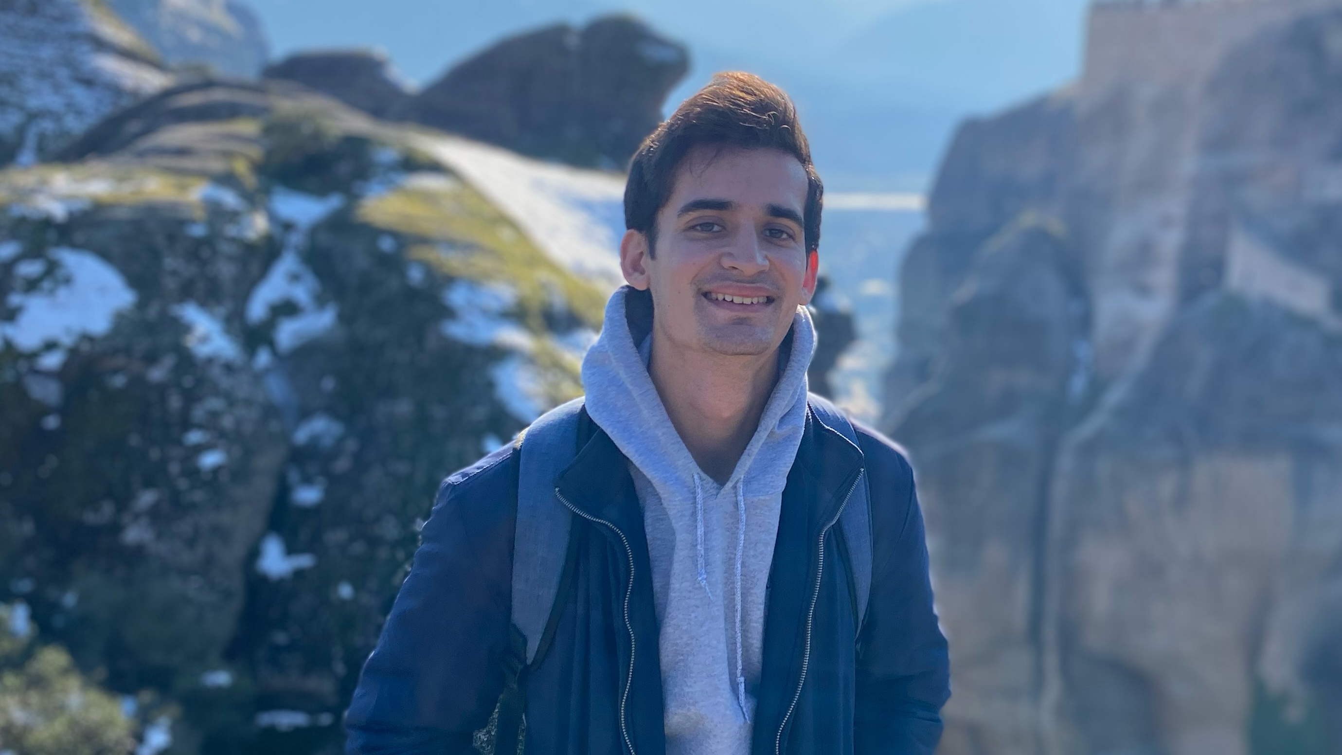 A man in a hoodie and jacket stands in front of a snow-covered mountainous landscape.