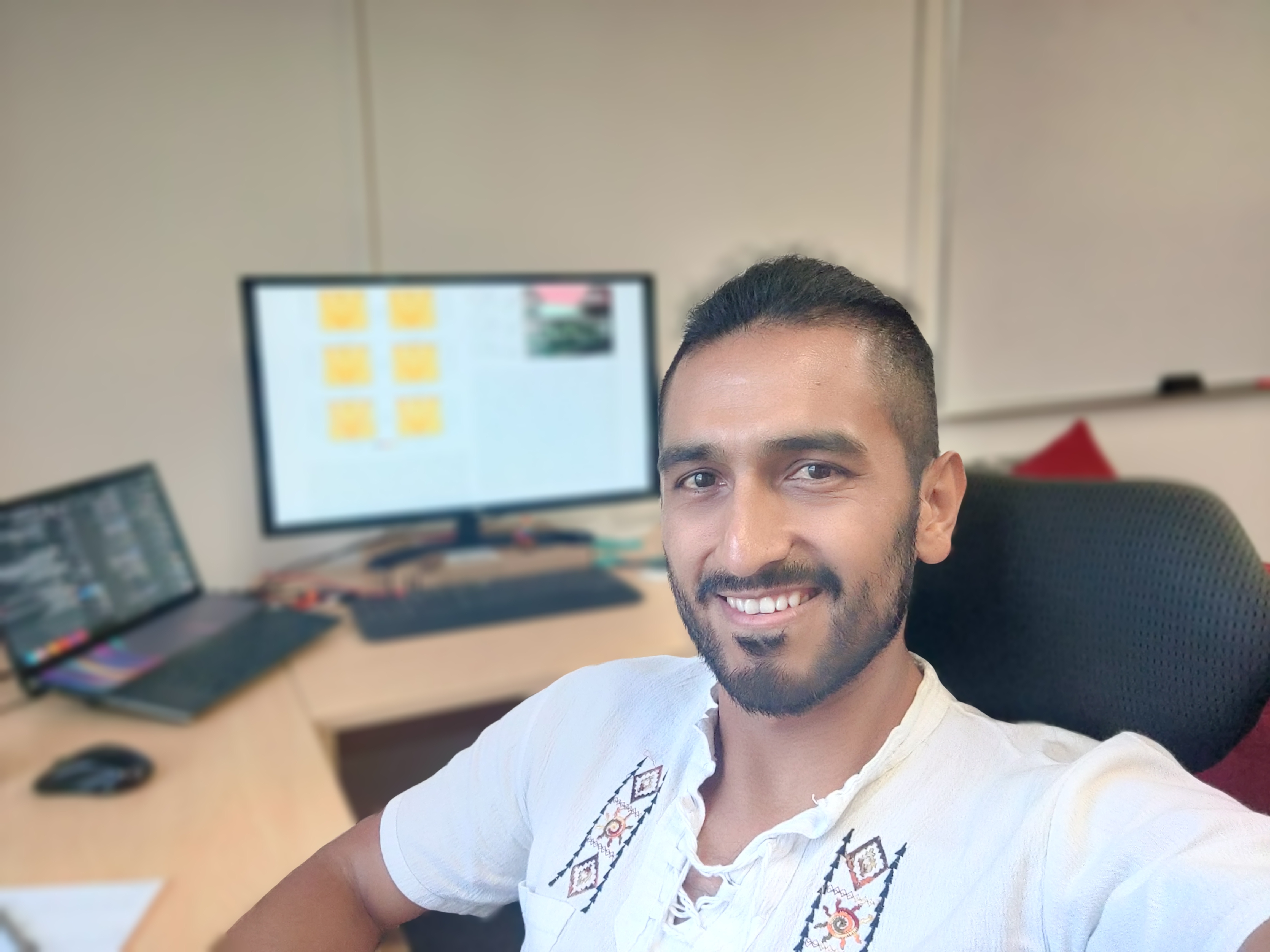 A smiling man with light brown skin sits in an office interior.