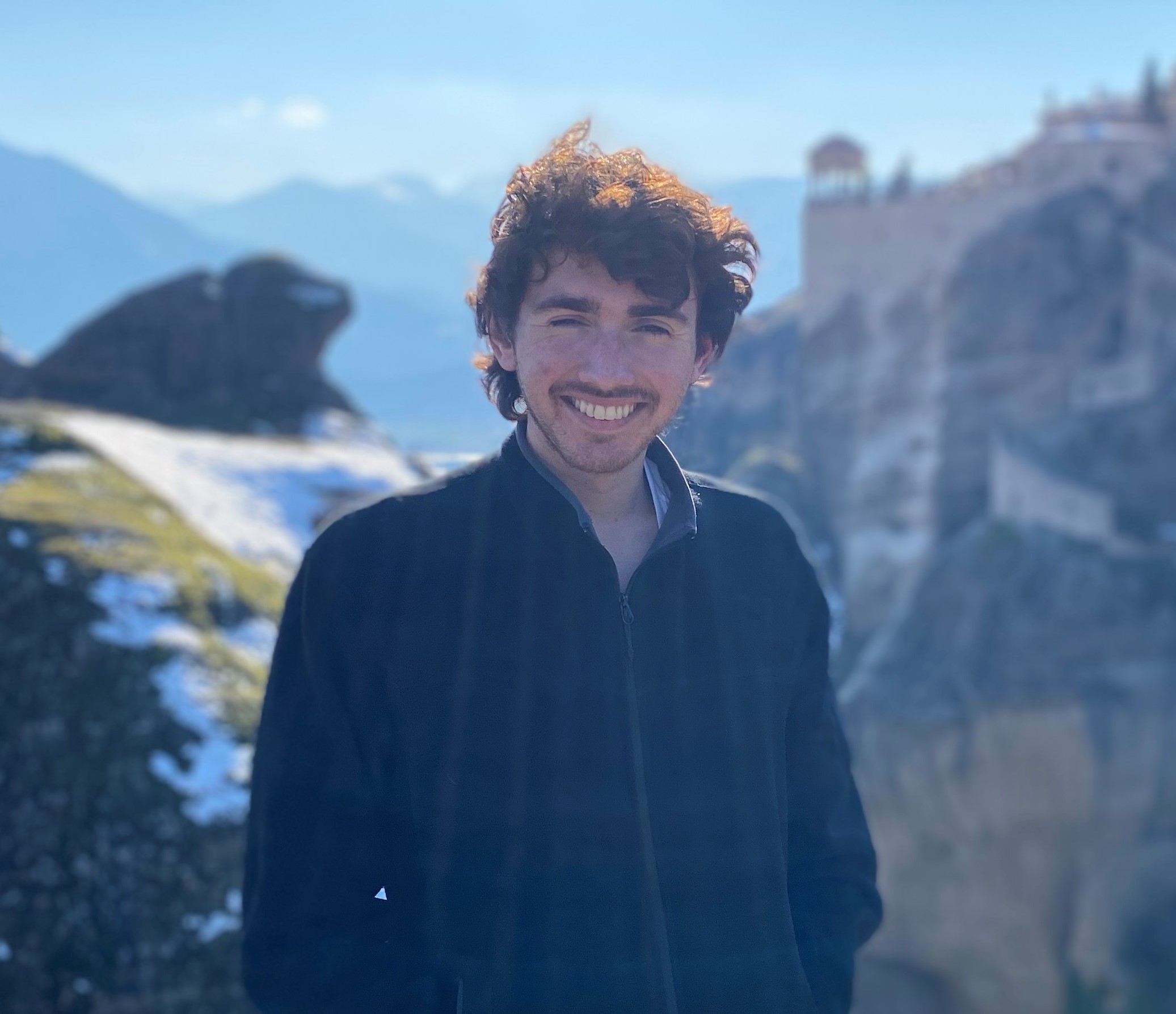 Smiling young man with light skin and dark hair appear in a wilderness setting with mountainous background.