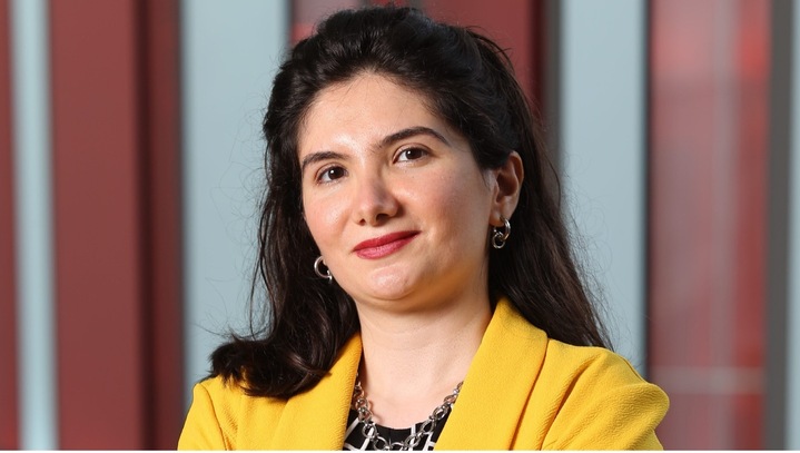 Zohreh Davoudi wearing a yellow jacket stands in front of red and white virticle stripes of varying widths.
