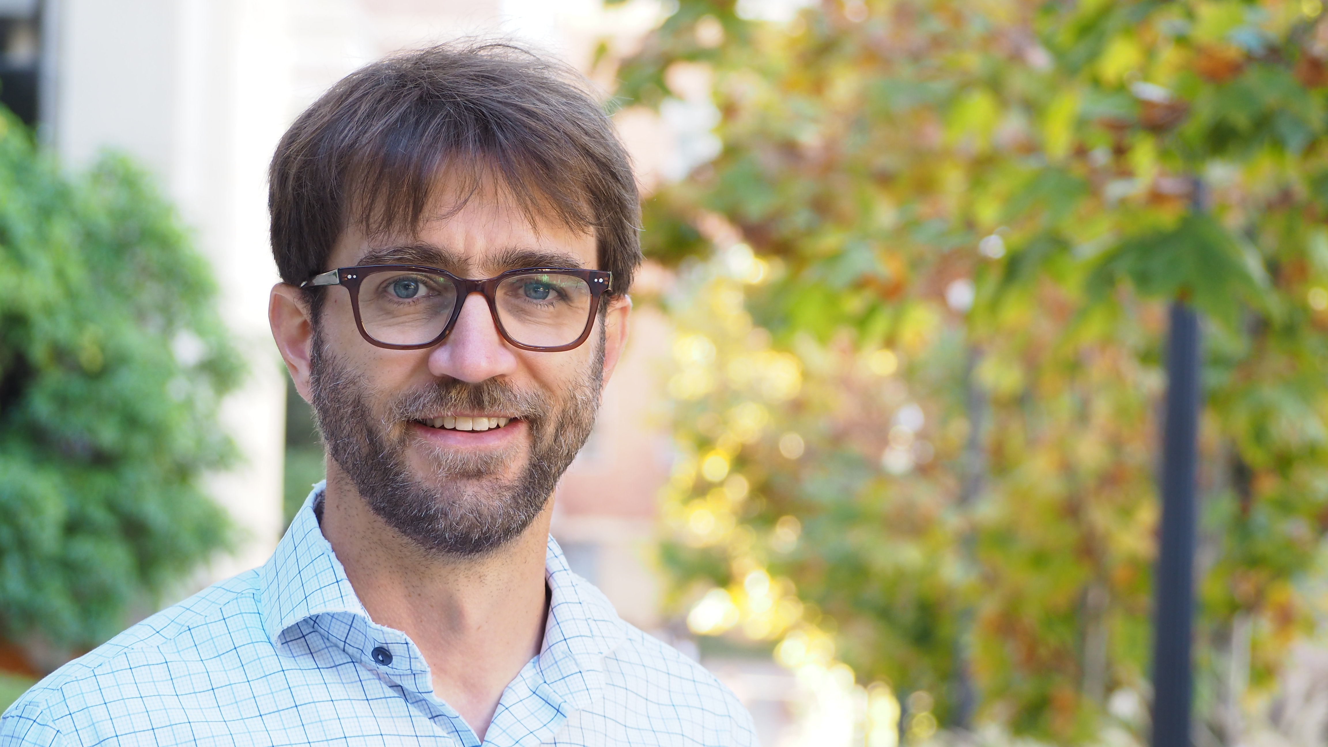 A man in a checkered shirt and glasses with out-of-focus trees in the background
