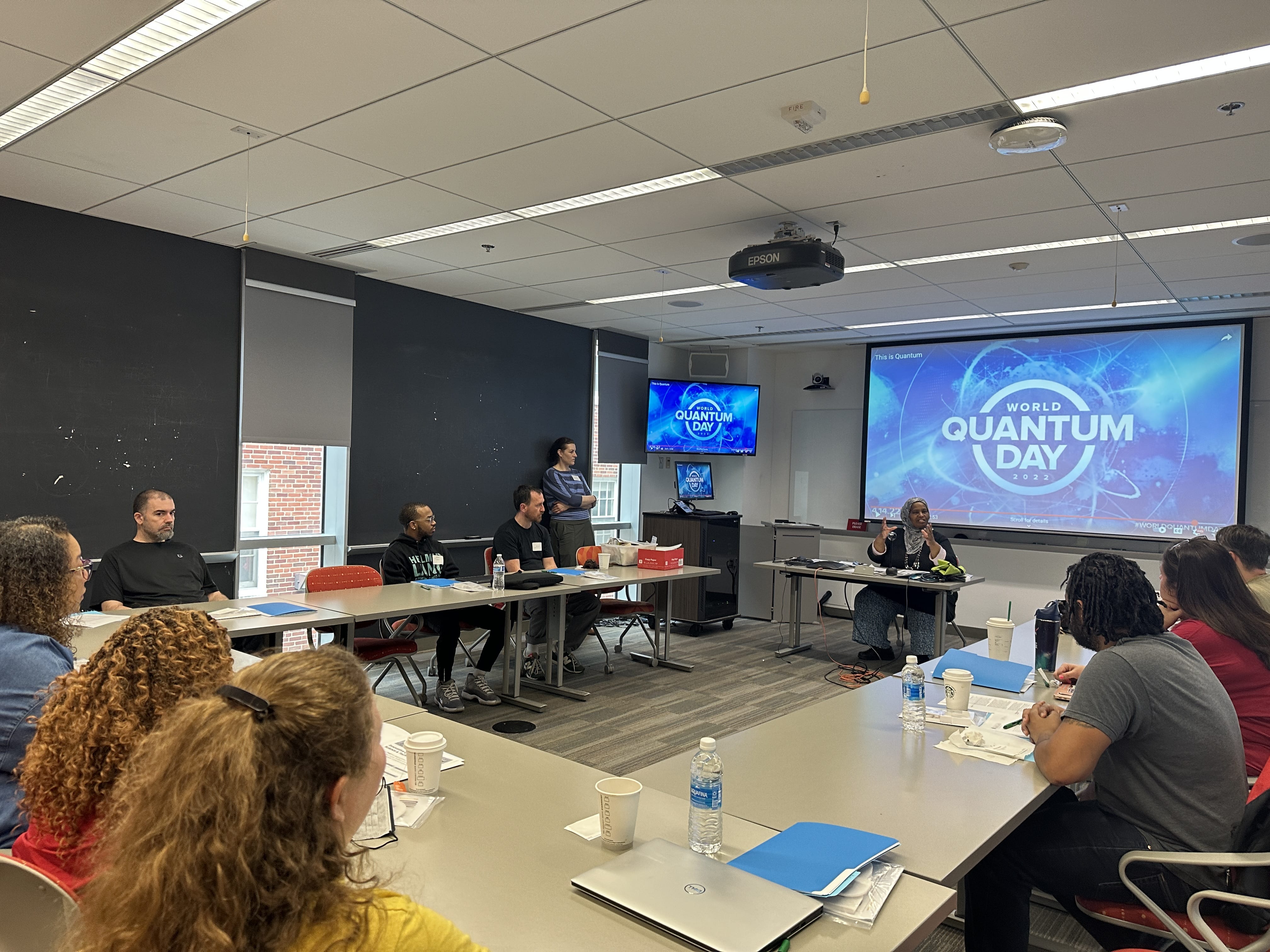 A group of educators and speakers participate in a World Quantum Day workshop inside a classroom, with presentation slides displayed on large screens.