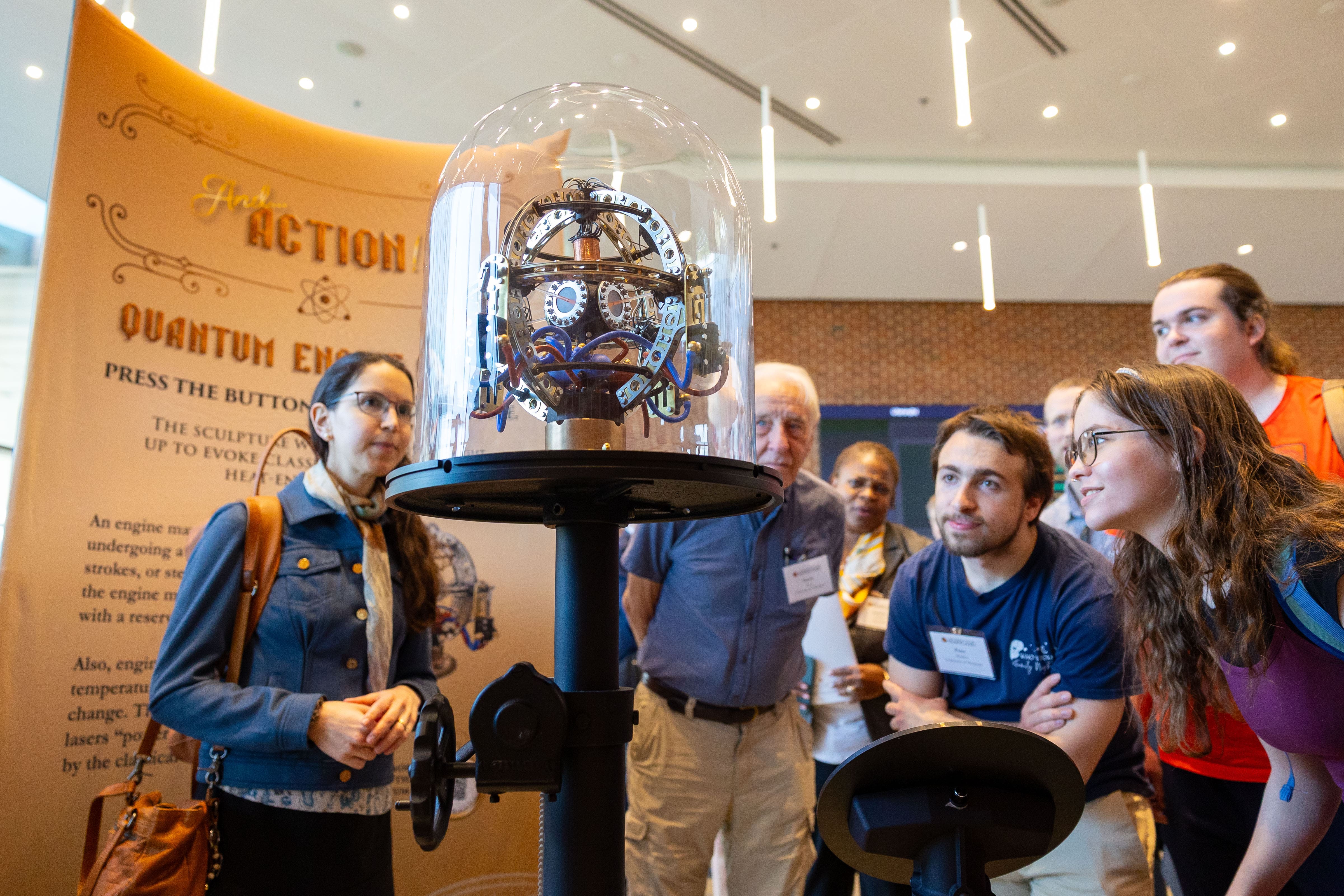 Visitors gather around a glass-encased quantum engine sculpture at a quantum science showcase, observing and interacting with the exhibit.
