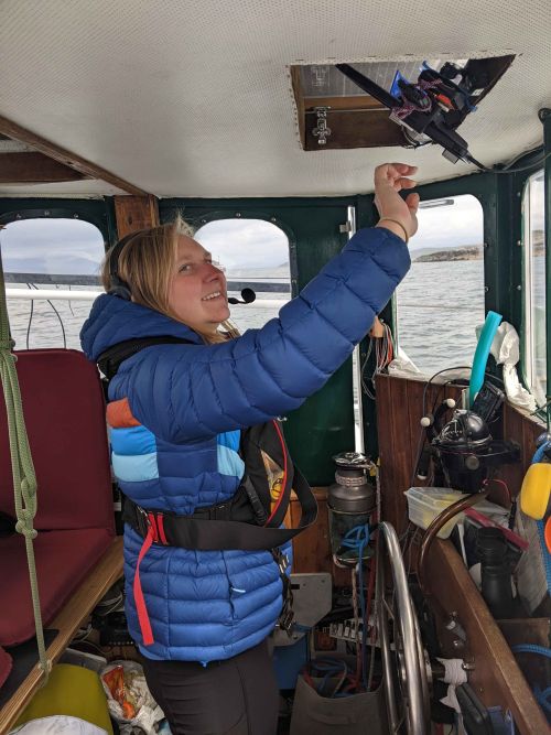 A woman in a blue jacket stands in a boat cockpit and is reaching up to controls hanging from the ceiling.